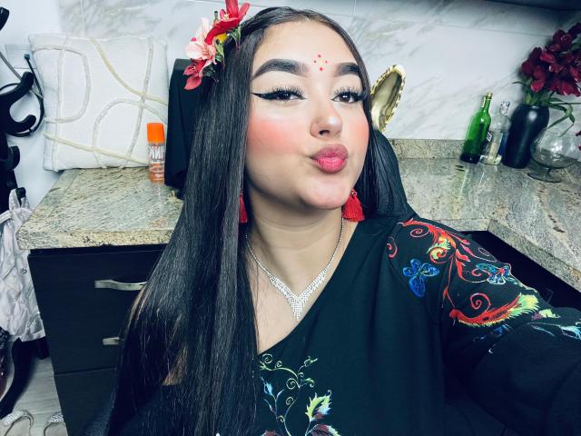 AnaShalimarr mit langem, schwarzem Haar, die ein schwarzes Kleid mit Blumenmuster, rote Ohrringe und eine rote Blume im Haar trägt, zieht ihre rosafarbenen Lippen zusammen. Ihr Make-up ist auffällig mit Eyeliner..