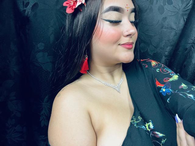 AnaShalimarr mit dunklem Haar, einer roten Blume, einem schwarzen Oberteil, farbenfroher Stickerei, roten Quastenohrringen, Eyeliner, rosa Rouge, einem Lächeln, einer silbernen Halskette, leicht freiliegenden Brüsten und einem Hintergrund mit einem schwarzen, blumigen Vorhang..