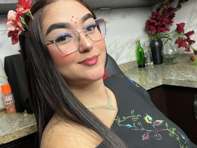 AnaShalimarr mit langen, schwarzen Haaren, Brille, einer roten Blume, Make-up-Punkten, pinkem Lippenstift, einem schwarzen Oberteil mit grüner Blumenstickerei, einer Halskette, lächelnd, mit einem Küchenk hintergrund, einer Flasche, Blumen und einer Arbeitsplatte..