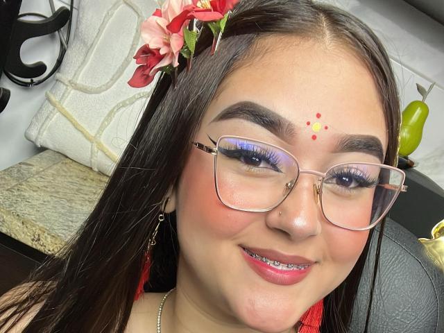 AnaShalimarr mit braunen Haaren, einer roten Blume, Brille, blauem Eyeliner, einem roten Punkt auf der Stirn, pinkem Lippenstift, einem Nasenpiercing, lächelnd, trägt rote Ohrringe und sitzt im Auto..