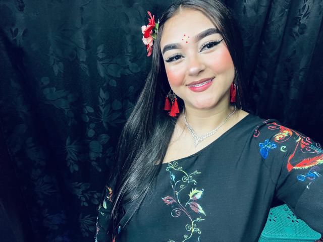 AnaShalimarr met lang zwart haar, rode franjes, een bloemenprint zwarte jurk, een rode bloem in haar haar, eyeliner en rode lipstick, lachend naar de camera, met een schattig gezicht en een kleine neus..
