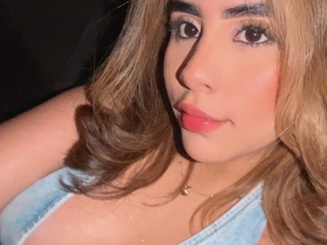 OlivaStellas has tan skin, pink lips, brown wavy hair, and blue eyes. She's wearing a light blue top that reveals her boobs. She's looking at the camera with a smile.