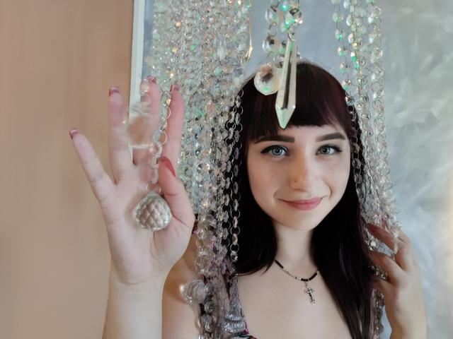NinaFlawlees with dark brown hair, holding a crystal-shaped ornament against a string of hanging crystals, smiling, wearing a necklace with a small key, showing her pink-manicured hand.