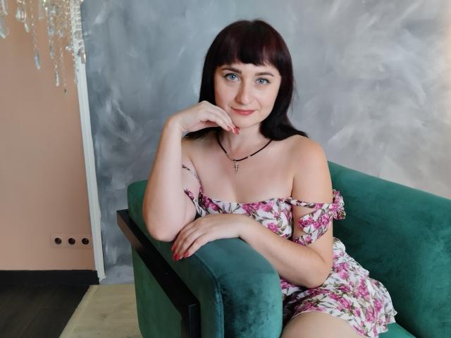 NinaFlawlees with red hair, fair skin, and blue eyes, sitting on a green couch, wearing an off-shoulder floral dress, hand on chin, smiling, black necklace with cross.