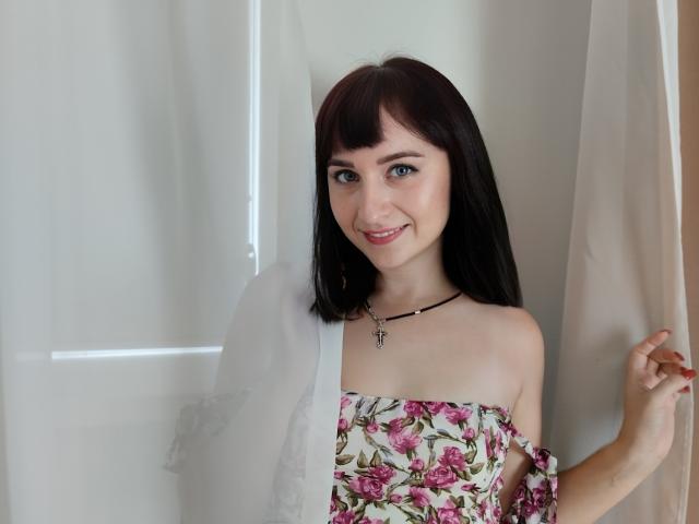 Nina, met donkerbruin haar, blauwe ogen en een roze bloemenjurk, lachend, terwijl ze een dunne gordijn vasthoudt, draagt een zwarte ketting met een kruis, heeft een lichte huid, is schattig en heeft kleine borsten..