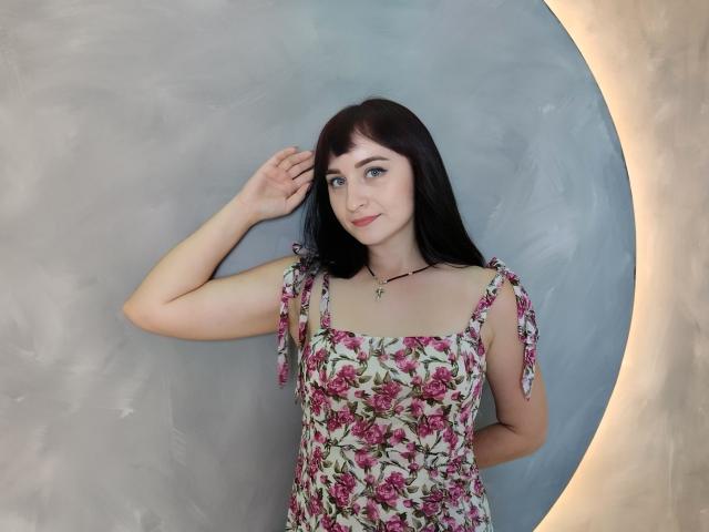 NinaFlawlees, con piel pálida, cabello negro, ojos azules, vestida con un vestido floral, con la mano levantada, sonriendo, con un collar, iluminación suave, de pie frente a una pared circular, gris y texturizada..