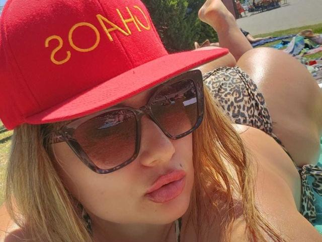 MirayDesire con un cappello rosso, la scritta "ZONAHO" in giallo, occhiali da sole tartarugati, labbra piene, capelli biondi, bikini a stampa leopardo, sdraiata su una spiaggia illuminata dal sole, con i glutei leggermente sollevati..