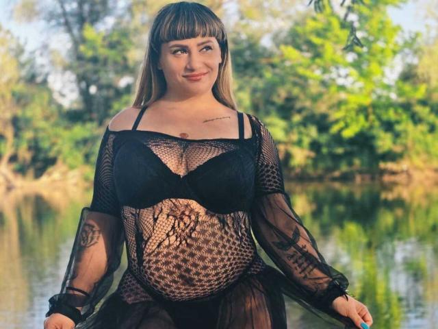 MirayDesire, con flequillo rubio, vistiendo una blusa de malla negra que deja ver sus grandes senos y un sujetador negro, de pie junto a un agua reflectante, sonriendo, con su vientre de embarazada visible..