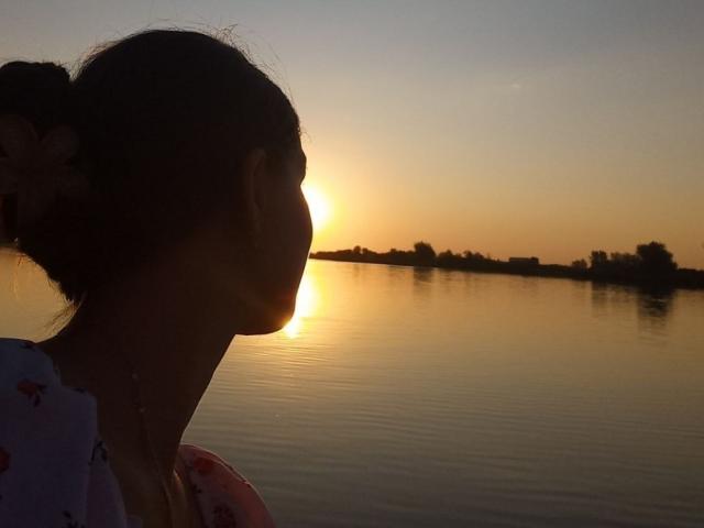 Silhouette of Didiene with her hair in a bun, facing right, showing her profile with visible neck and a floral dress, at sunset with the sun near water's edge.