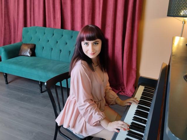 NinaFlawlees with red bangs, sitting on a chair, playing a piano in a pink dress, green velvet sofa, and red curtains in the background. Boobs not visible.