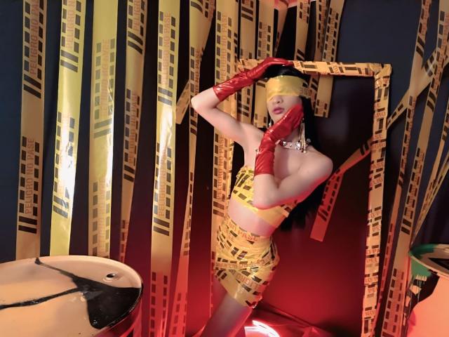 KendallSky in yellow latex outfit, red gloves, and yellow mask, standing with hands on head, showing small tits, yellow patterned background, and a reflective surface on the left.