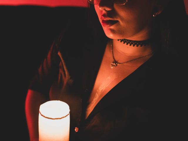 LiliethWolf with a black choker and necklace, wearing a low-cut shirt, holding a glowing candle, red light on her face, partially visible lips, and glasses.