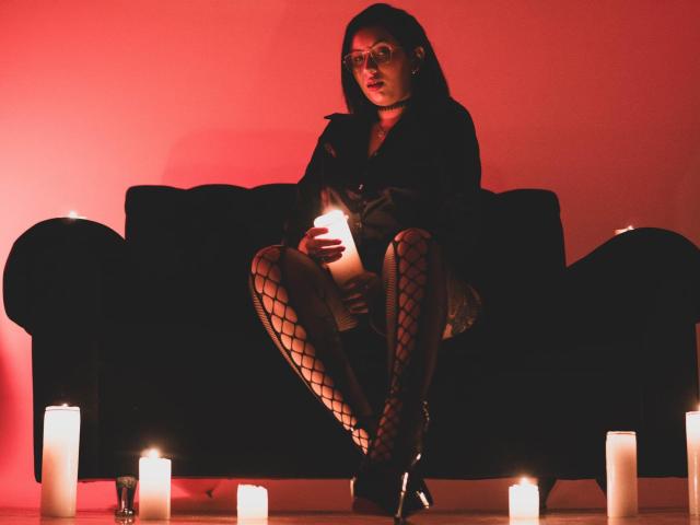 LiliethWolf with glasses, dark hair, black top, fishnet stockings, holding candle, sitting on black couch, surrounded by candles, red light, looking down, serious expression.