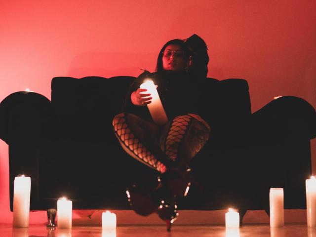 LiliethWolf sitting on a black couch, holding a candle, wearing fishnet stockings and black boots, surrounded by lit candles, in a red-lit room. Her ass is prominently displayed.