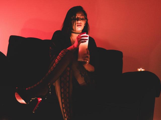 LiliethWolf with red glasses, black choker, fishnet legs, high heels, holding paper, sitting on black couch, red lighting, sexy, looking down, dark hair, seductive pose.