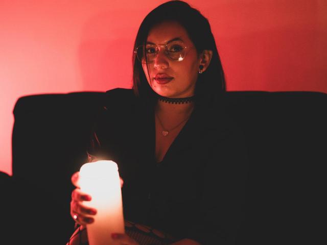 LiliethWolf with red-lit face, holding a glowing candle, wearing glasses, choker, and black outfit, sitting on a black couch, looking at camera.