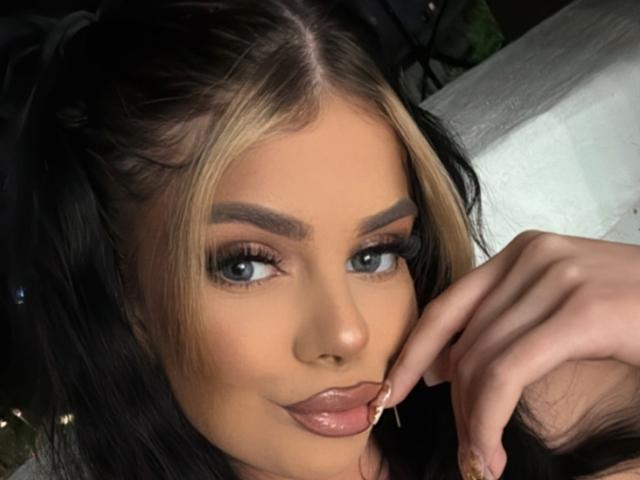 IvyRaven has blonde and black hair, blue eyes, and is sucking her glossy lips with her finger near them. Her eyebrows are thick, and she has long, dark lashes.