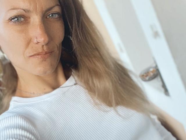DaneeSoHot, mit einem Nasenring, trägt ein weißes Rippoberteil, hat langes, braunes Haar und einen ernsten Gesichtsausdruck. Auf dem Selfie sind ihre kleinen Brüste und ein leichtes Schmollen zu sehen..