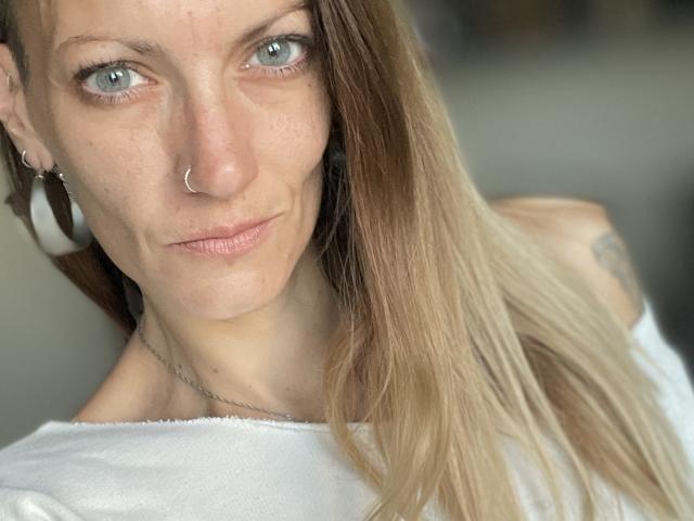 DaneeSoHot hat helgrüne Augen, einen Nasenring und langes blondes Haar. Sie trägt ein weißes Oberteil, hat ein leichtes Lächeln und ein Tattoo an ihrer rechten Schulter..