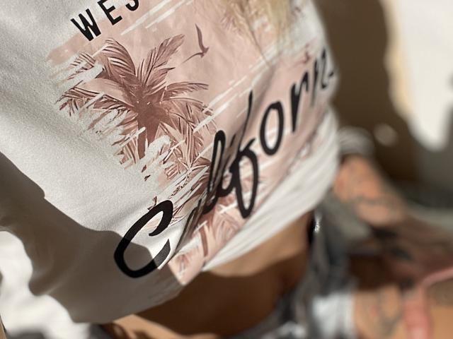A camiseta branca da DaneeSoHot, com a estampa de palmeira e a inscrição "California", cobre parcialmente seus seios, com uma tatuagem iluminada pelo sol no braço direito e um fundo desfocado..