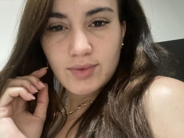 AnitaLux has light brown skin, long brown hair, and a slight smile. She's wearing a gold necklace, has small earrings, and her hand is near her face.