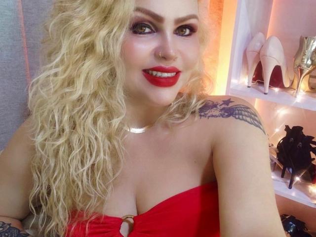 AnnaRichie with curly blonde hair, red lipstick, nose ring, and tattoo on right shoulder, wearing a red strapless top, showing her tits, smiling at the camera.