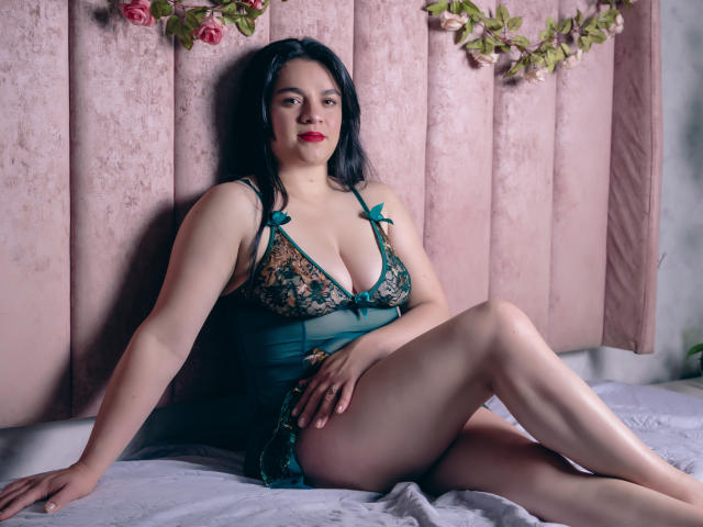 DannaBlake with black hair, wearing a green lace lingerie with teal bows, sitting on a bed, legs crossed, displaying her tits and pussy, pink wall background, flowers.