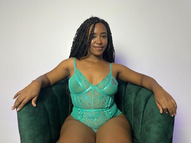 NaomiReyes, dark-skinned, with long braids, wearing a green lace lingerie bodysuit, sits in a green velvet chair, arms resting on arms, smiling slightly, with small, perky tits.
