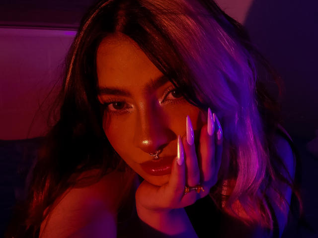 WillowRosse with long wavy hair, glowing pink and purple lights, nose ring, shiny pink lips, and long white nails touching her face, looking seductively at the camera.