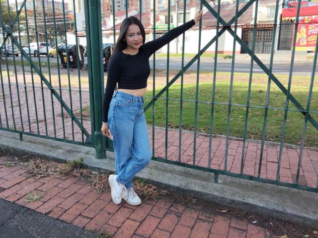 TinyLaurent with medium tits, black crop top, high-waisted blue jeans, and white sneakers, leaning on green fence, arm raised, standing on red brick sidewalk, day, smiling.
