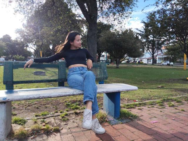 TinyLaurent sits on a blue-green bench, wearing a black long-sleeve top and blue jeans, with white sneakers, in a park with green grass and trees.