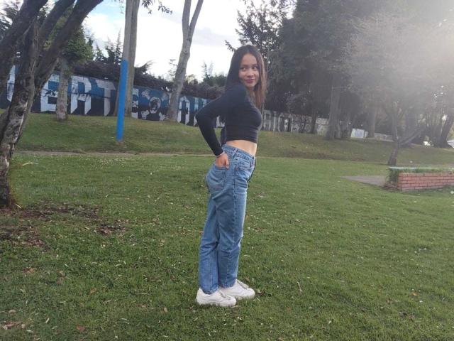 TinyLaurent stands on grass with hand on hip, wearing a black long-sleeve top, blue jeans, and white sneakers, showing her ass and smiling at the camera.