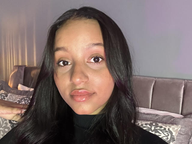 RaquelZafd has medium brown skin, black straight hair, brown eyes, and glossy pink lips. She's wearing a black turtleneck, with a grey headboard and pillows in the background.