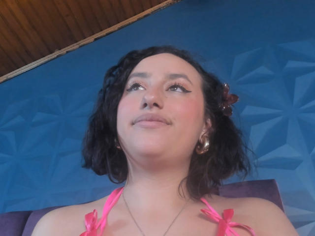 JudyCampbells has curly black hair, pink halter top, hoop earrings, and a subtle smile, looking up with eyeliner accentuating her eyes. Her small nose and slight blush are visible.