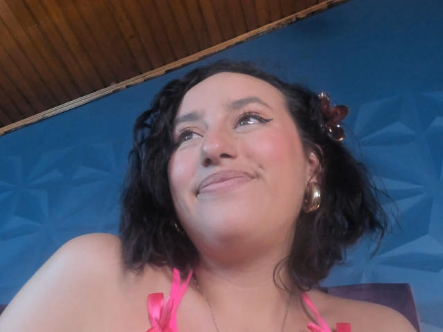 JudyCampbells has curly black hair, pink eyeliner, and a small nose ring, smiling with a slight smirk, wearing pink straps and silver earrings, against a blue geometric wall.