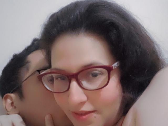 CoupleFantasticErotic with dark hair, red glasses, and a slight smile, resting head on another's shoulder. Both have light skin. The second person is mostly out of frame.