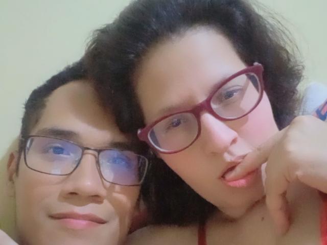 CoupleFantasticErotic with dark curly hair, red glasses, and a nose ring, leans on CoupleFantasticErotic with short black hair and glasses, both smiling closely.