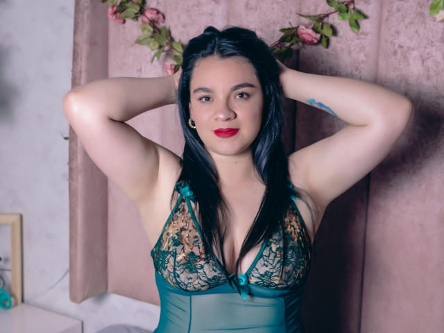 DannaBlake with black hair, hands in her hair, wearing a green lace lingerie top, showing her tits, red lipstick, and a tattoo on her right arm.