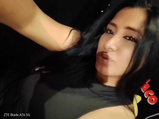 MariaCatalina with black hair, brown eyes, and a slight pout, wearing a black top, having her hair pulled by a hand on the left. Her face is centered.