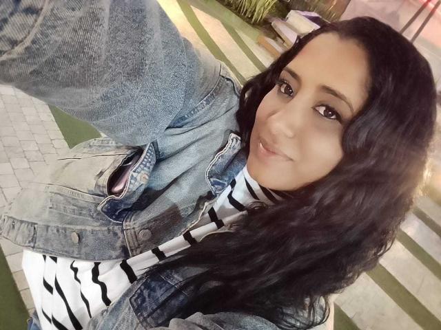 MariaCatalina with wavy black hair, brown eyes, and light skin, wearing a denim jacket and striped white turtleneck, smiling at the camera. Her tits are partially visible.