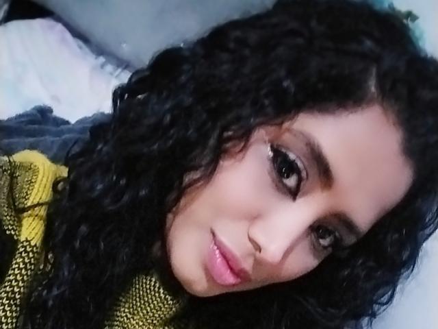 MariaCatalina has curly black hair, dark eyes, and pink lips. She's wearing a yellow-green knit top, looking at the camera with a slight smile. Her nose is pierced.