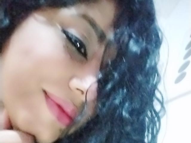 MariaCatalina's face with curly black hair, dark eyeliner, and pink lipstick, smiling slightly, with a cute nose and soft skin. Her black eyes are slightly open.