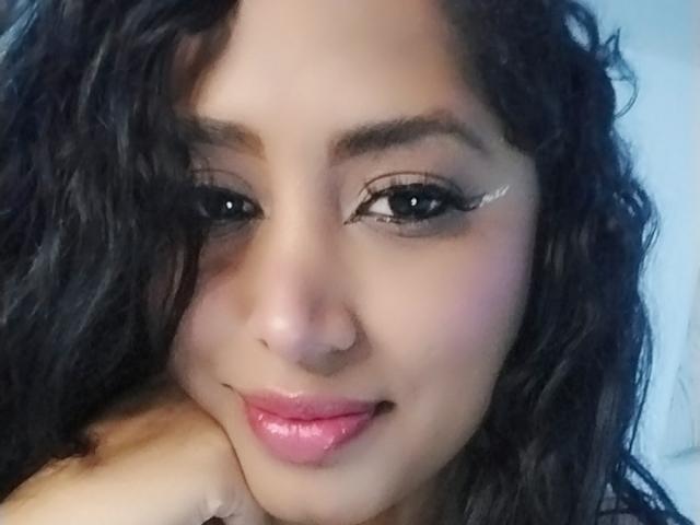 MariaCatalina has curly black hair, brown eyes with white eyeliner, and glossy pink lips, resting her head on her hand. Her nose is pierced.