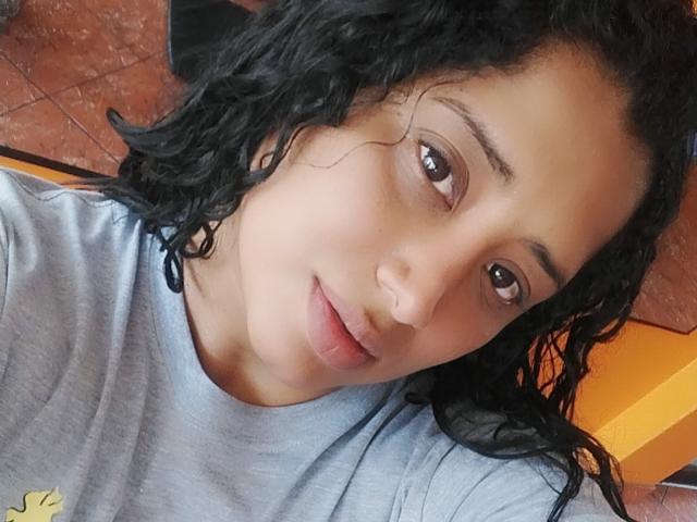 MariaCatalina with curly black hair, brown eyes, and light brown skin, wearing a gray shirt, smiling slightly, and looking at the camera. Her nose is small and her lips are full.