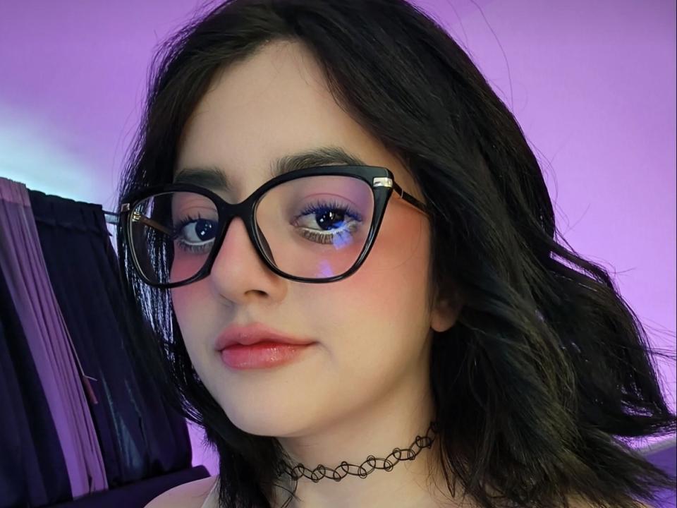 KatheGarcia with black glasses, pinkish-blue eye reflection, pinkish blush on cheeks, black choker, and wavy dark hair, looking slightly to the right with a neutral expression.