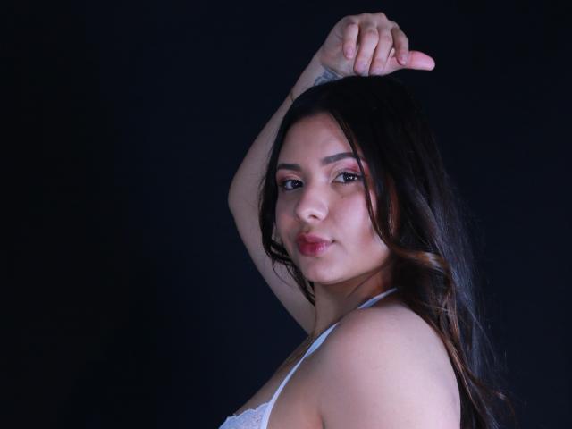 OrianaGarciax with long dark hair, white lace bra, arms raised, looking over shoulder, slightly pouty lips, dark background, tattoo on left arm, soft light.