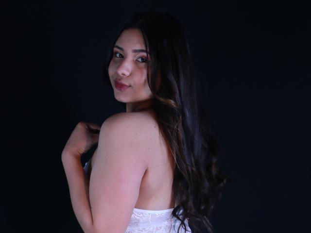 OrianaGarciax with long dark hair, smiling at camera, white lace top, bare shoulders, holding top with right hand, looking over shoulder, black background.