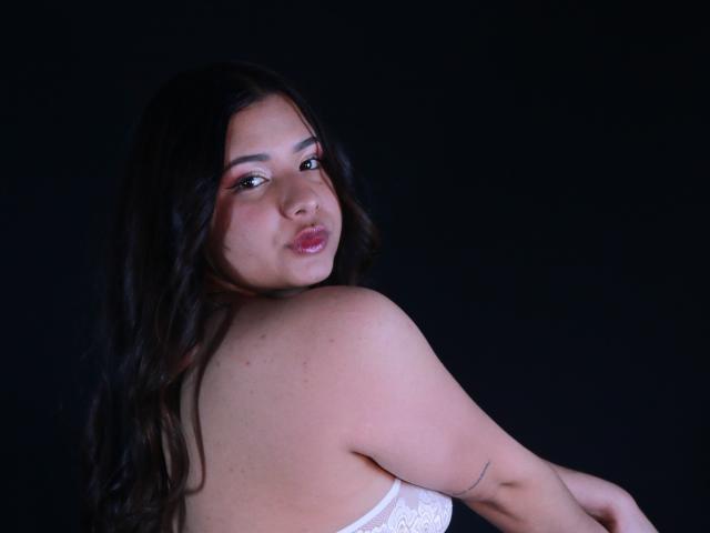 OrianaGarciax with long dark hair, pink eyeshadow, and white lace top, looking over her shoulder, showing her back and a small tattoo on her right arm.