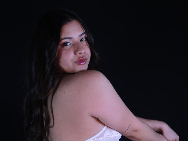 OrianaGarciax with dark wavy hair, looking back, has pink eye makeup, white lace bra, and a small tattoo on her upper arm, against a dark background.