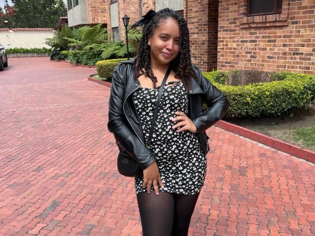 ArtemisaS stands in a red-bricked driveway, wearing a black floral dress, black leather jacket, and black tights. She has curly black hair, a black bow, and a hand on her hip.